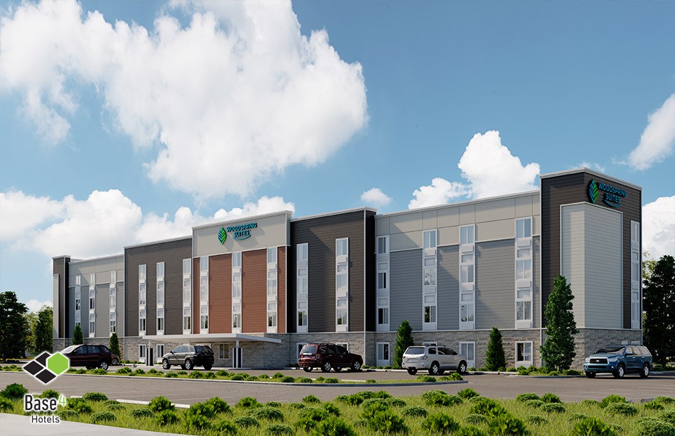 Modern WoodSpring Suites hotel showcasing a long and sleek architectural design with a light gray facade, accented by the brand's signage, set in a lush green landscape designed by BASE4.